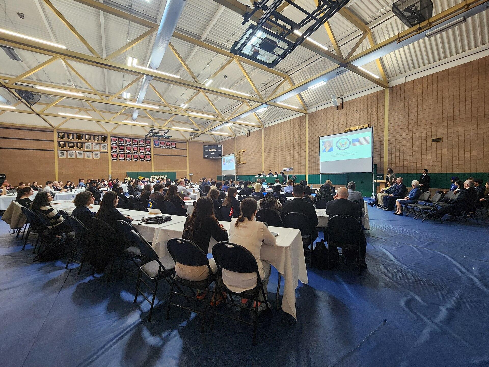 Students and adults seated at conference tables during a Model UN general assembly session in a school gymnasium